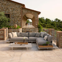L-shaped outdoor sofa in soft gray fabric with beige cushions, set against a stone backdrop and lush greenery.