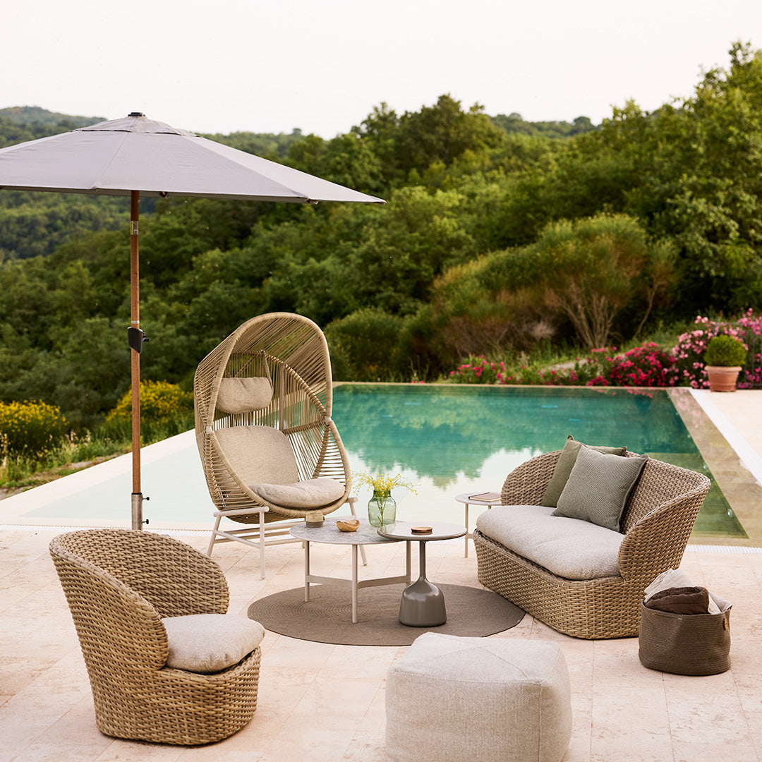 "Neutral beige patio furniture with soft gray cushions and a round taupe rug, surrounded by greenery and a blue pool."