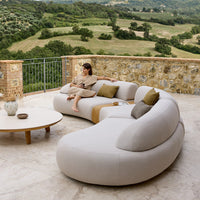 Curved beige outdoor sofa with muted green and yellow accent pillows, set against a natural landscape backdrop.