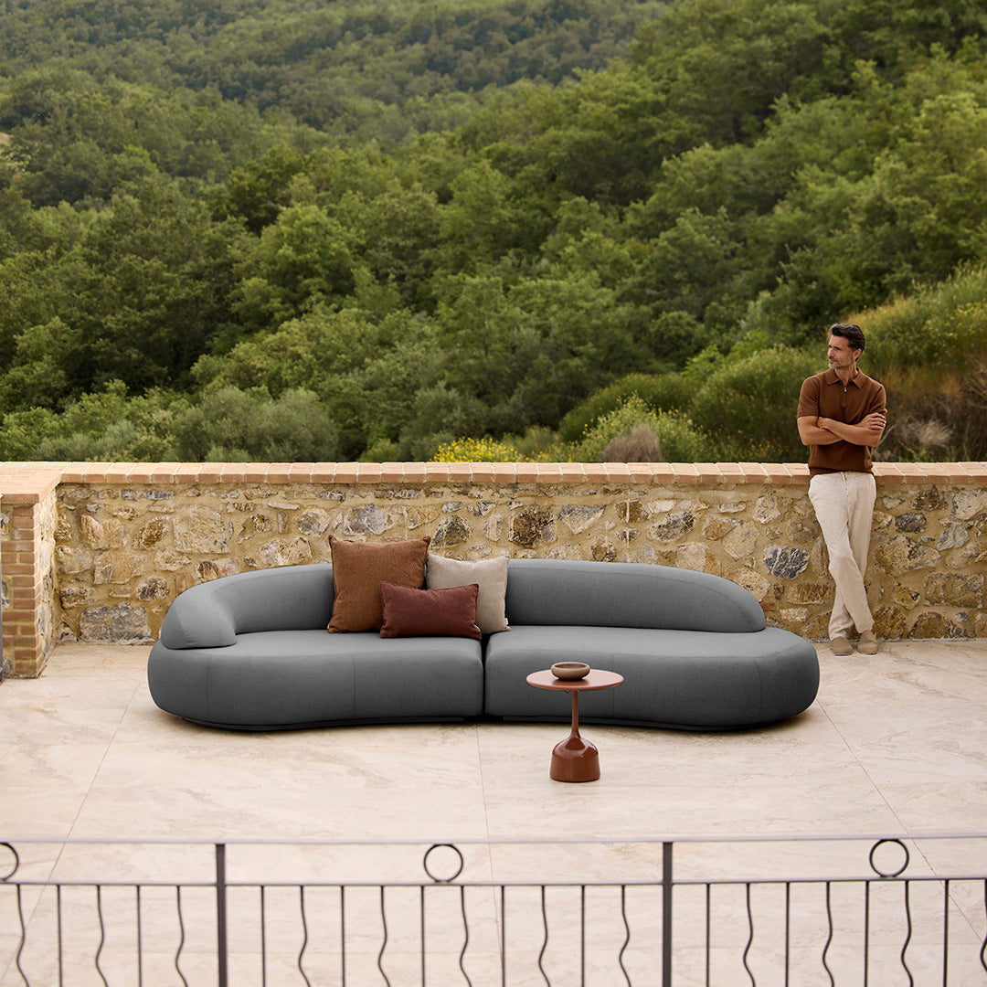Curved gray sofa with cream and brown accent pillows, set against a natural green landscape.