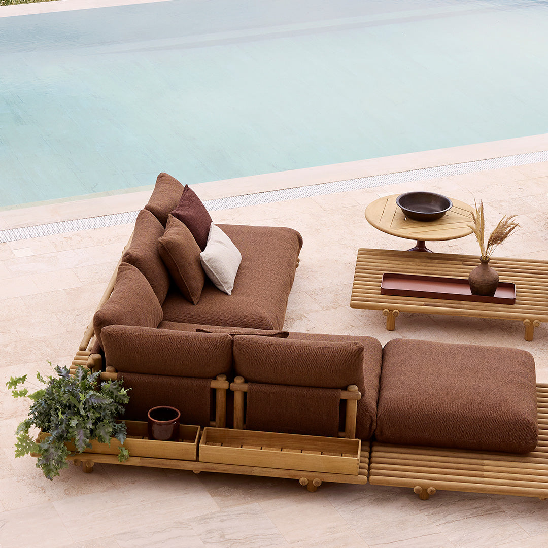 Brown sectional sofa with cream and burgundy pillows set on a wooden platform beside a pool, featuring minimalist decor.