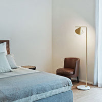 Elegant gold floor lamp beside a brown chair, set against soft beige and gray bedroom decor, enhancing modern aesthetics.