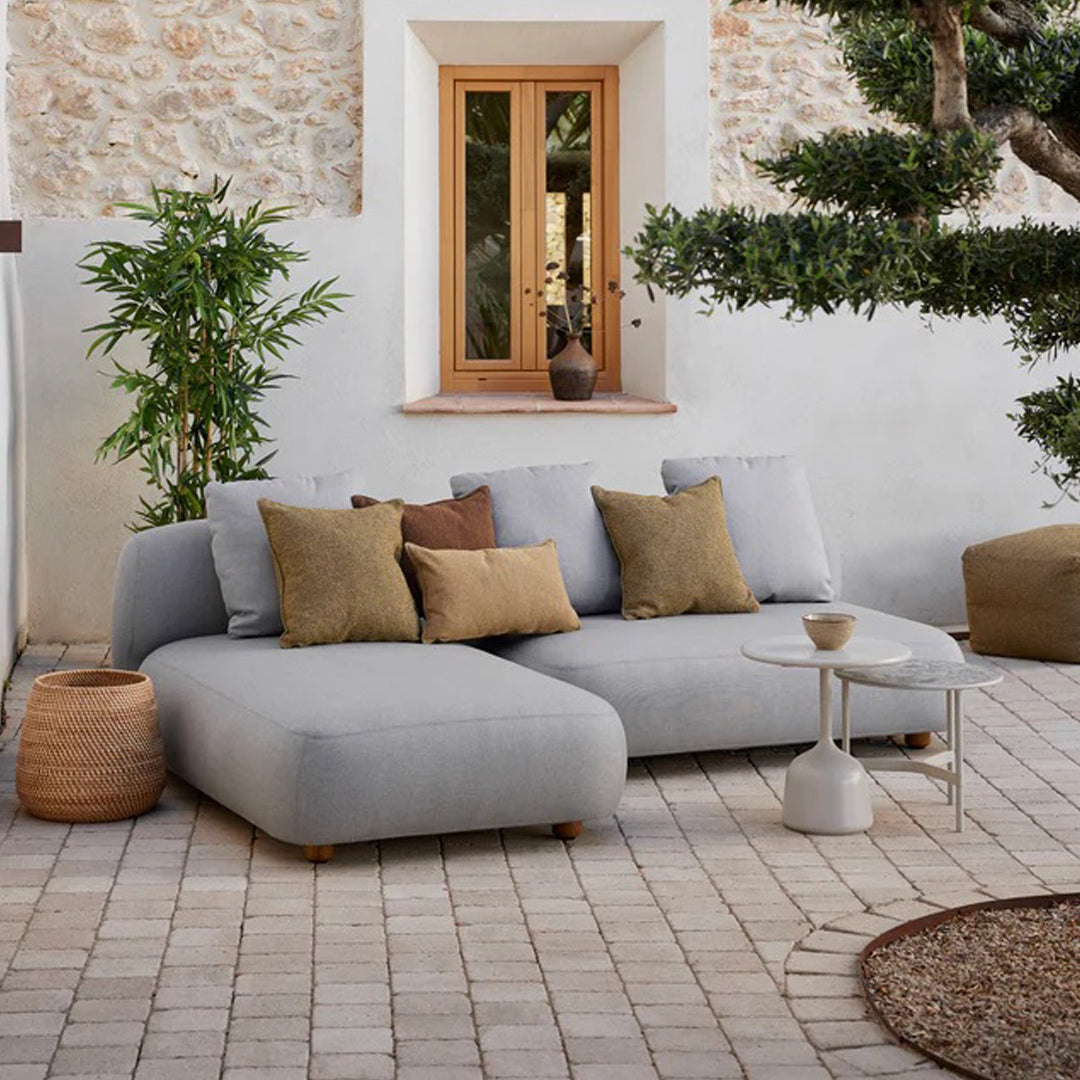 Light gray sectional sofa with brown and olive green pillows, set against a rustic stone background and natural elements.