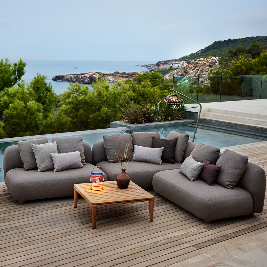 Gray outdoor sectional sofa with mixed-patterned cushions and a natural wood coffee table, set by a scenic coastal view.
