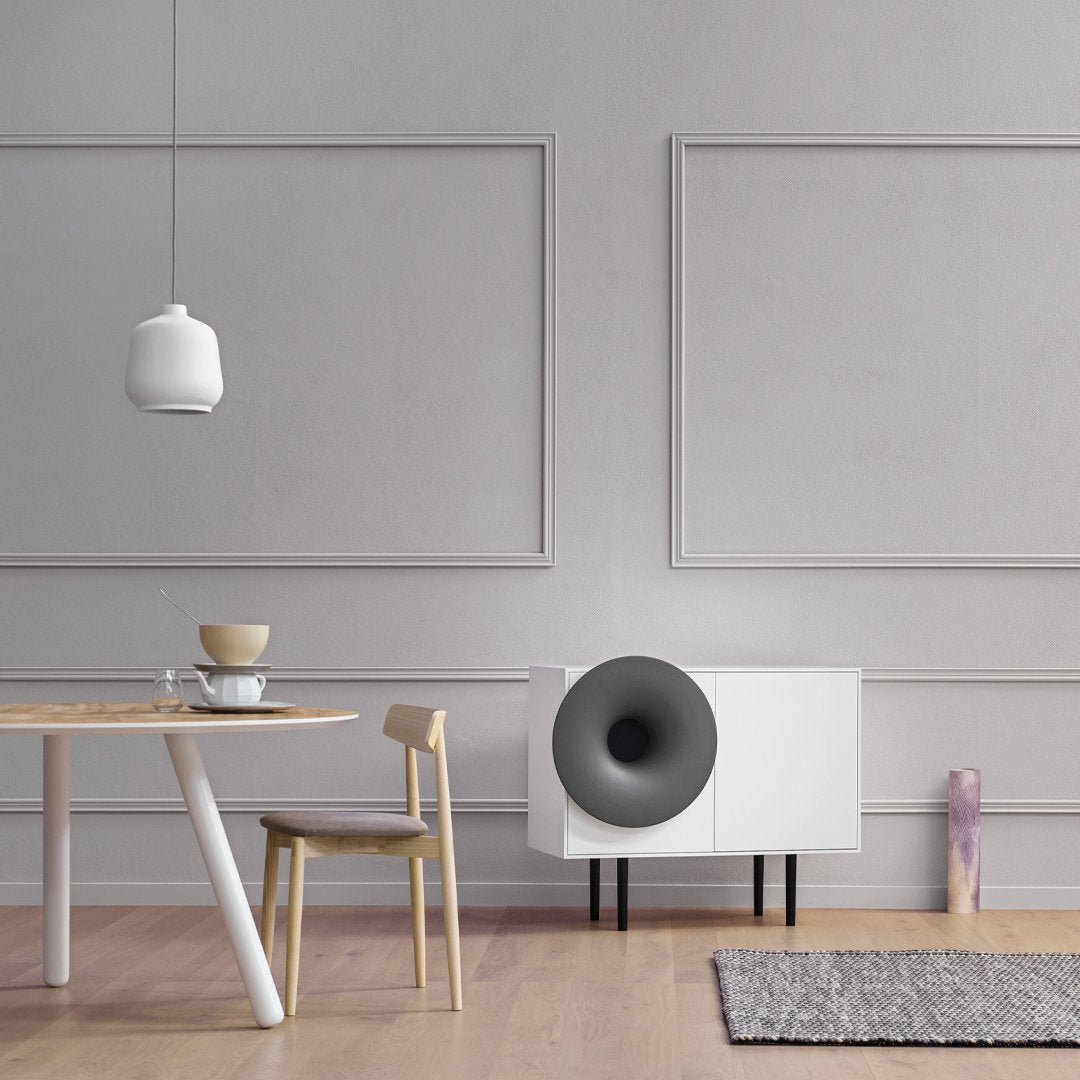 Modern white cabinet with black speaker, gray walls, wooden table, and light-colored natural wood chair in a minimalist setting.