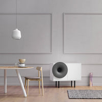Modern white cabinet with black speaker, gray walls, wooden table, and light-colored natural wood chair in a minimalist setting.