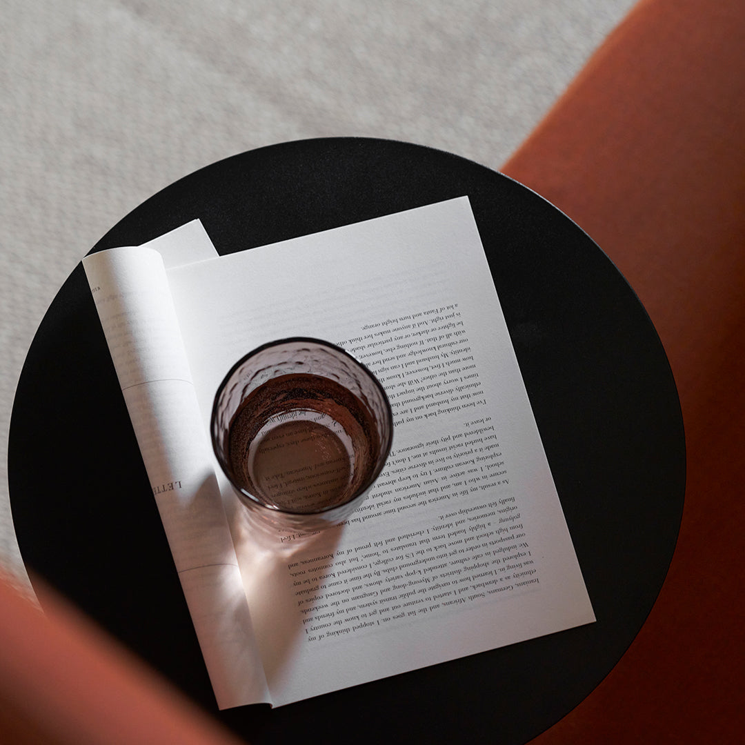 Black round table featuring a clear glass and an open white book with minimalist text on light gray texture background.