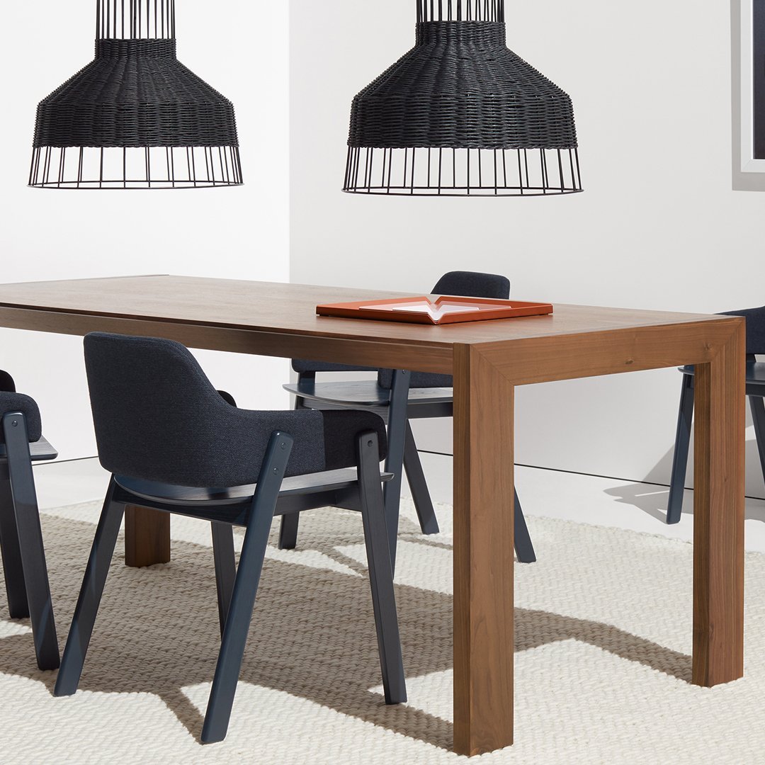 Wooden dining table in rich brown with black chairs and geometric black pendant lights. Neutral textured rug underneath.