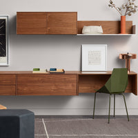 Modern wooden desk in warm brown tones with green chair, textured gray rug, and minimalist white shelving.
