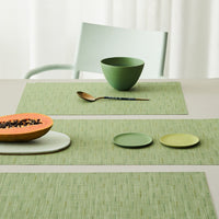 Green textured placemats with circular dishes in soft green and orange bowl, styled on a light table setting.