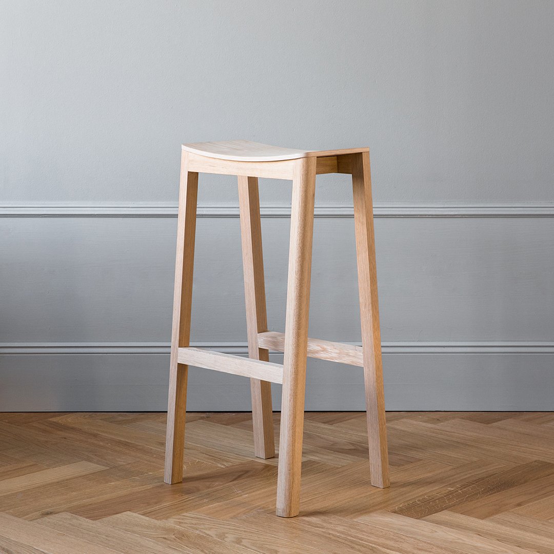 Light wood bar stool with a smooth, natural finish against a textured gray wall and herringbone wood floor.