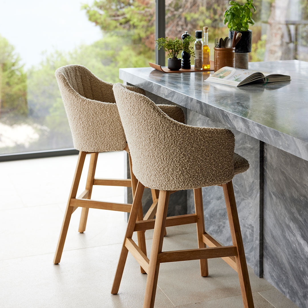 Neutral beige upholstered bar stools with textured fabric and wooden legs against a modern marble countertop.
