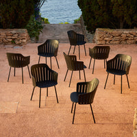 Set of eight modern black slatted chairs arranged outdoors on a stone patio, surrounded by greenery and water.
