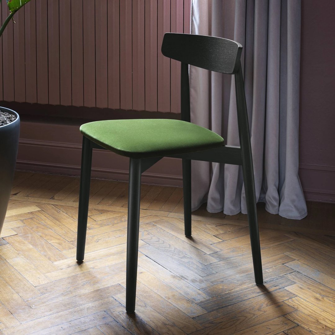 Green upholstered chair with a sleek black frame on a herringbone wooden floor, complemented by soft gray curtains.