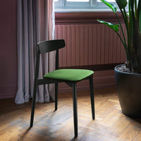 Black wooden chair with a vibrant green seat, set against a pastel pink wall and natural wood floor.