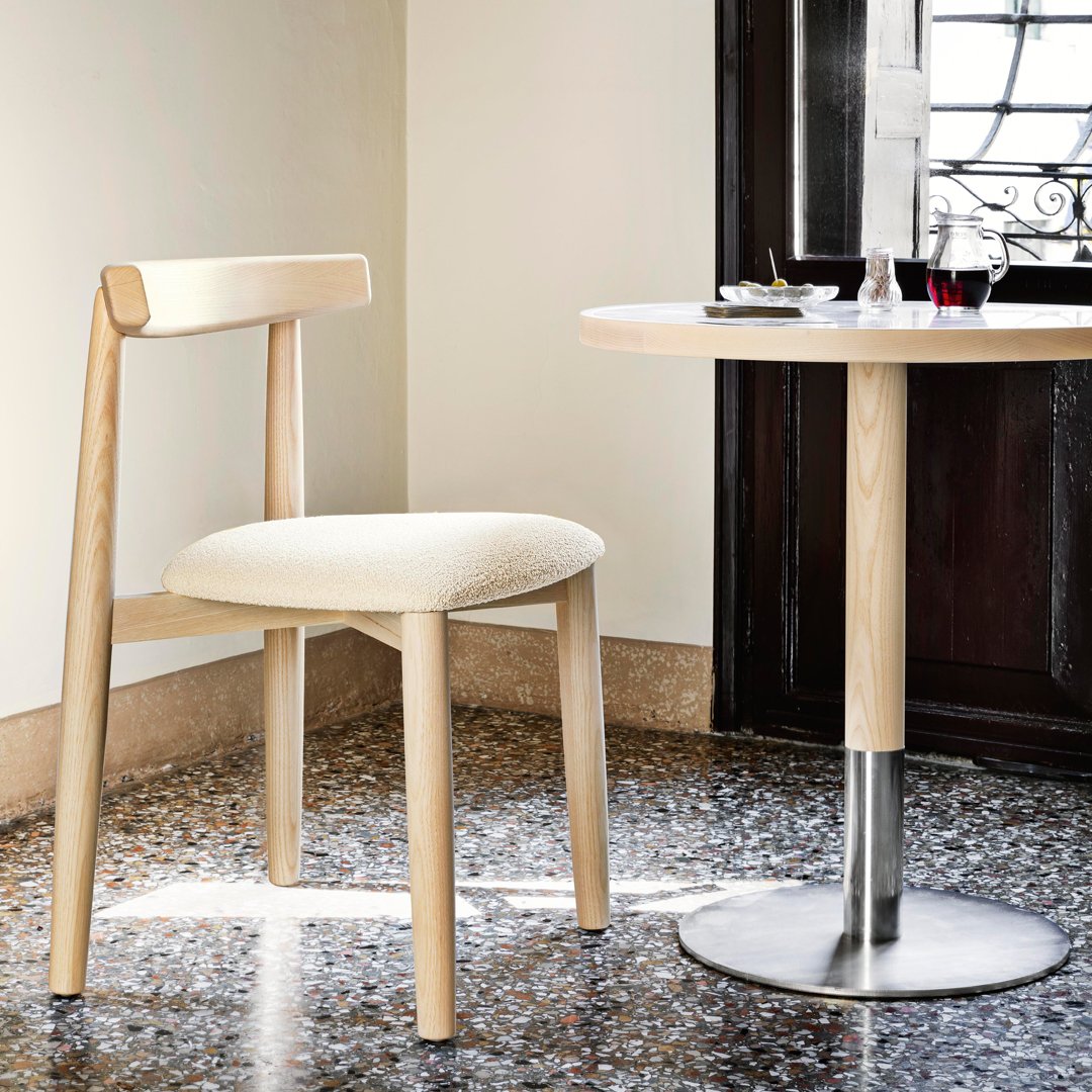 Light wood chair with a cream cushion against a neutral wall and round silver table, featuring a classic minimalist design.