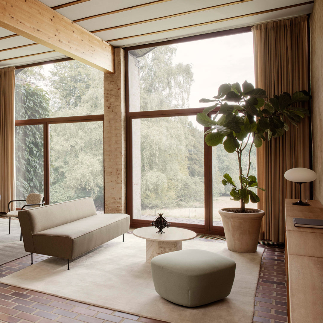 Modern living room with neutral tones, soft beige sofa, green ottoman, and large windows showcasing natural greenery.