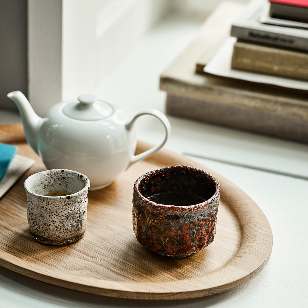 Ceramic teapot and cups with speckled earthy tones on a wooden tray, featuring warm textures and subtle patterns.