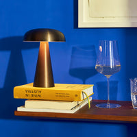 Bronze table lamp on yellow book stack against vibrant blue wall, with clear glassware nearby. Modern, sleek design.