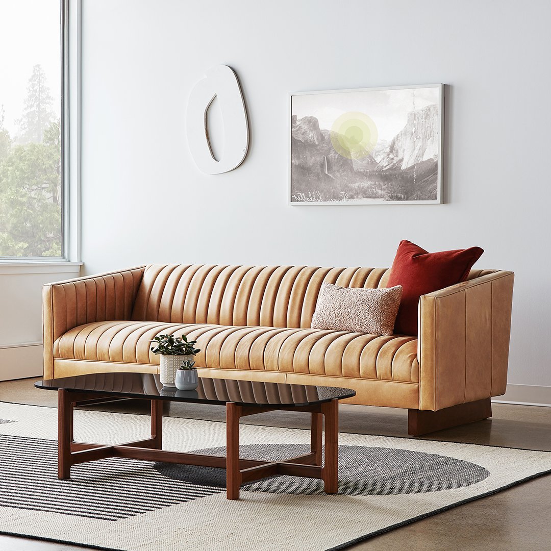 Light tan ribbed leather sofa with a rich red and neutral throw pillows, paired with a dark wood coffee table and patterned rug.