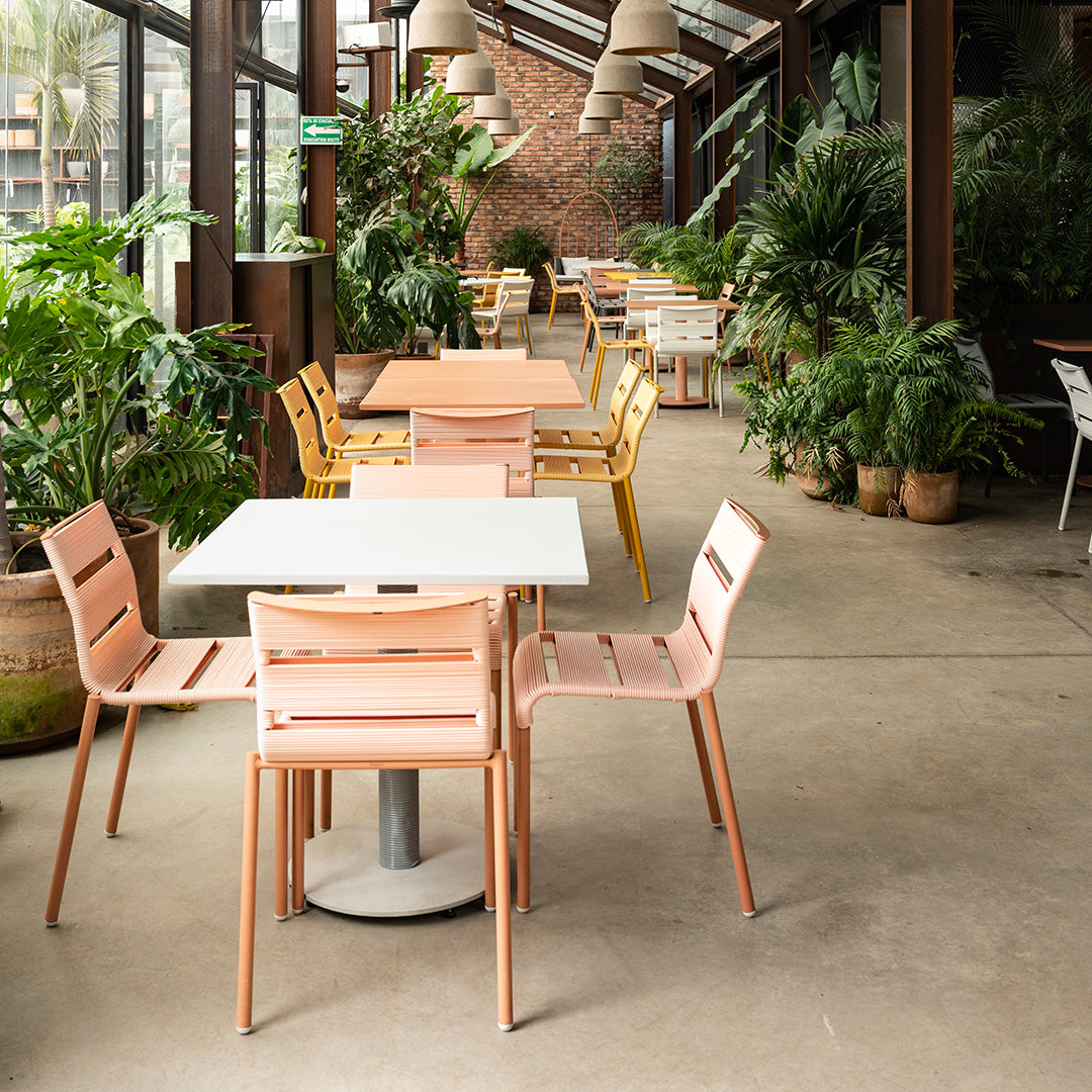 Modern cafe with pastel pink and yellow chairs, white tables, and lush greenery in natural light.