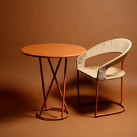 Rust-orange round table with a woven beige chair on a warm brown background. Modern design with contrasting colors.