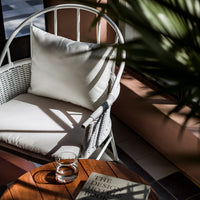 White woven chair with a neutral cushion, wooden table, and soft shadows from nearby greenery. Modern, minimalist aesthetic.