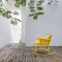 Bright yellow rocking chair with intricate woven pattern, set against a minimalist white background and earthy brick flooring.
