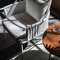 White wicker chair with a soft cream cushion, set against dark tiles and a warm wooden table. Subtle shadows and highlights.