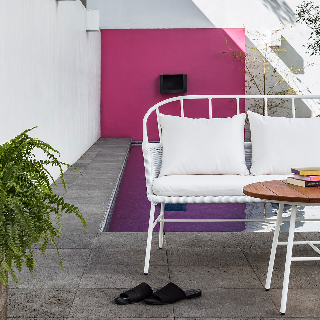 White outdoor sofa with soft cushions, contrasting against a vibrant pink wall and gray stone patio. Elegant modern decor.
