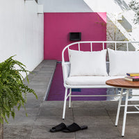 White outdoor sofa with soft cushions, contrasting against a vibrant pink wall and gray stone patio. Elegant modern decor.