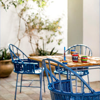 Blue wicker chairs and a wooden table, accented with colorful striped bowls, set against a backdrop of greenery.