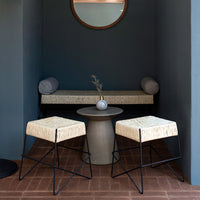 "Neutral beige woven stools with black frames, set against a deep blue wall and terracotta floor."