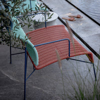 Colorful patio chair with a coral red woven seat and contrasting light mint green arms, on a stylish outdoor setting.