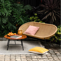 "Natural wicker chair with a pink pillow, yellow patterned cushion, and a wooden table surrounded by lush greenery."