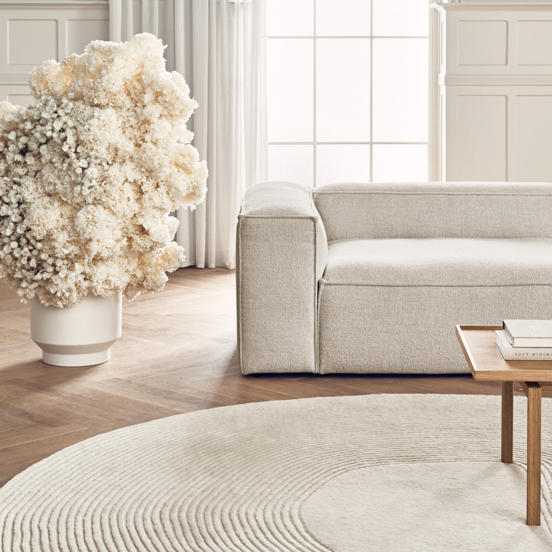 Neutral-toned sofa with a textured fabric, paired with a circular cream rug and soft, muted floral arrangement.