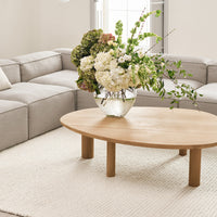 Natural wood coffee table with a round shape, surrounded by soft gray furniture and vibrant green floral arrangements.