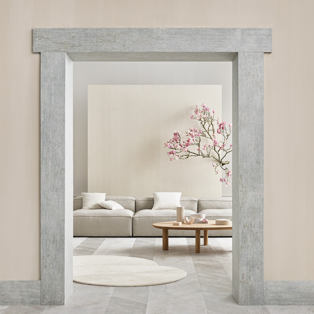 "Minimalist living room with soft beige walls, gray stone archway, lush pink floral accents, and a round white rug."