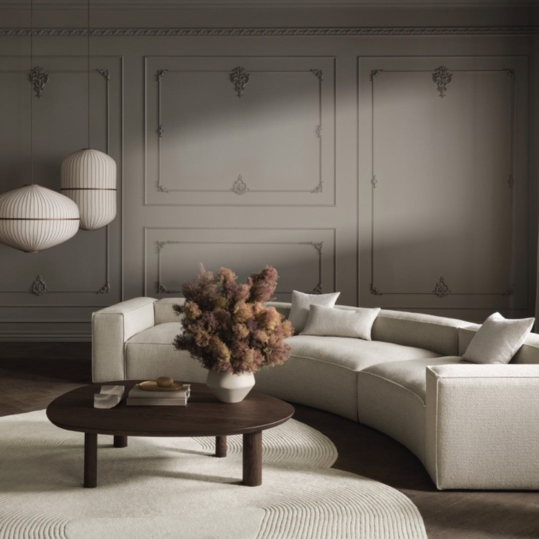 Neutral-toned curved sofa with textured fabric, surrounded by warm pink florals and elegant light fixtures against a gray wall.