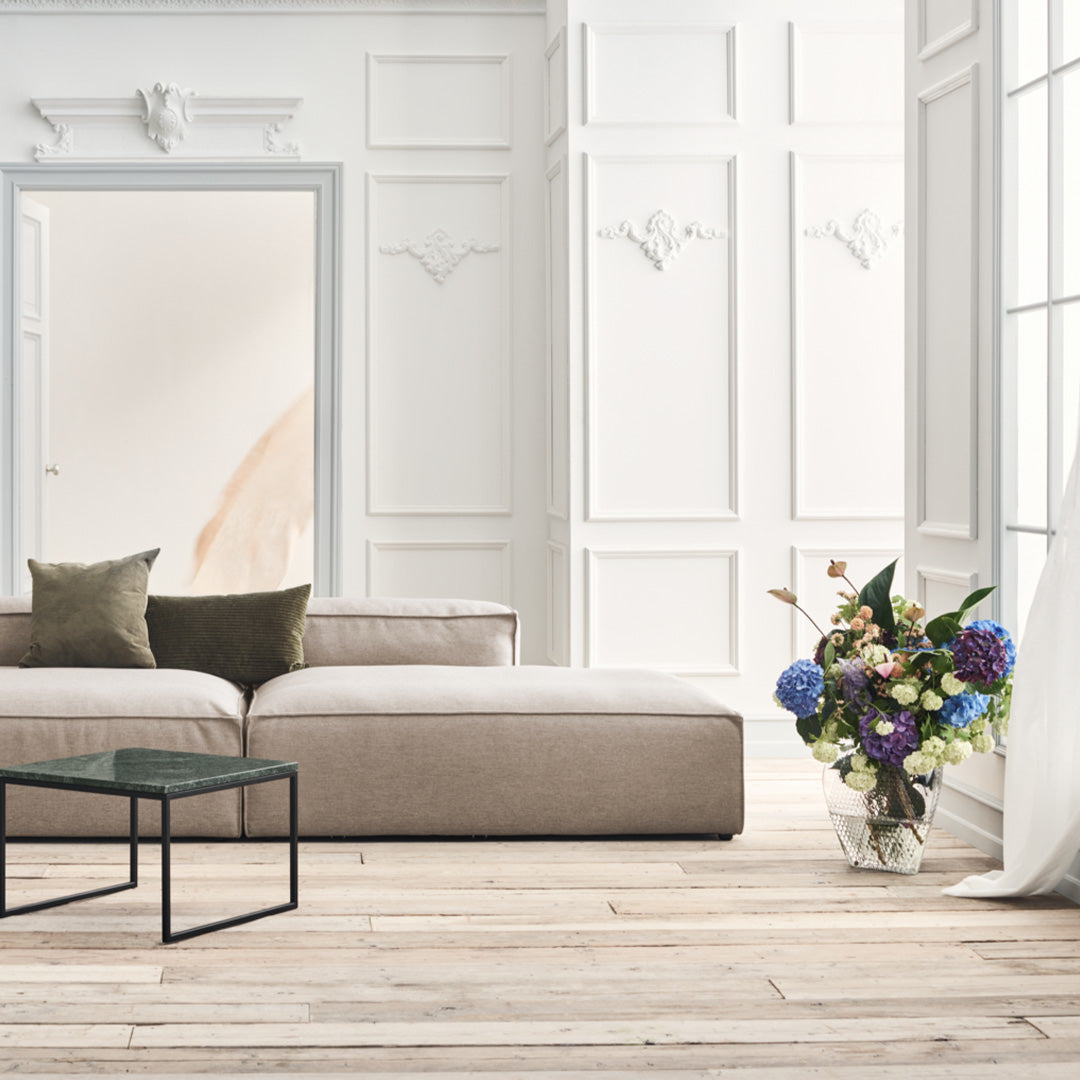 Beige sectional sofa with green and brown cushions, set against white walls and wood flooring, accented by colorful floral arrangement.