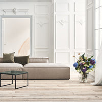 Beige sectional sofa with green and brown cushions, set against white walls and wood flooring, accented by colorful floral arrangement.