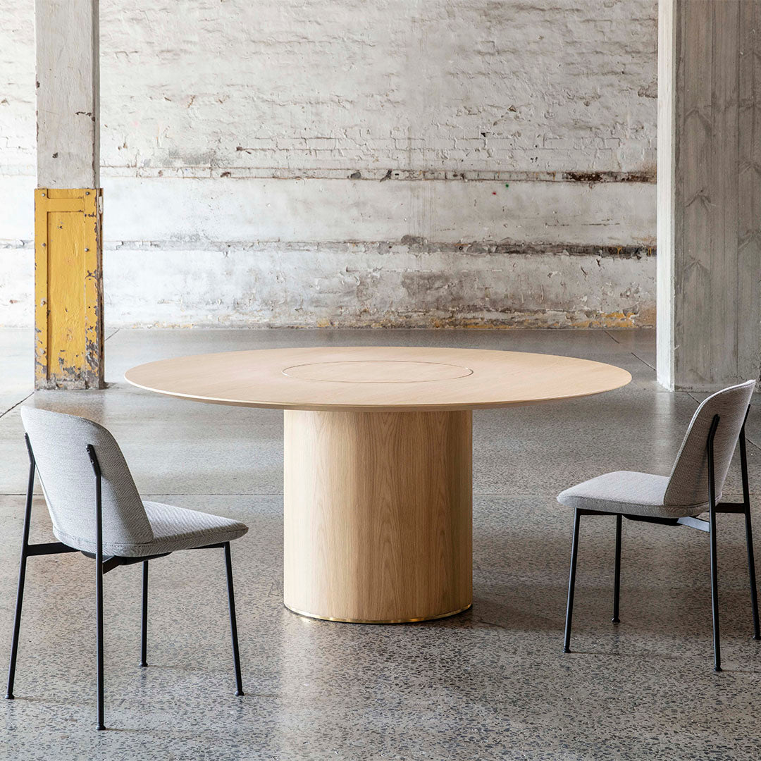Light oak round table with a smooth finish, paired with soft gray upholstered chairs against a minimalist backdrop.