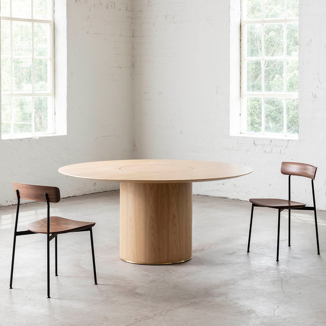 "Round wooden table with a light oak finish and two dark wood chairs featuring black metal legs in a bright room."