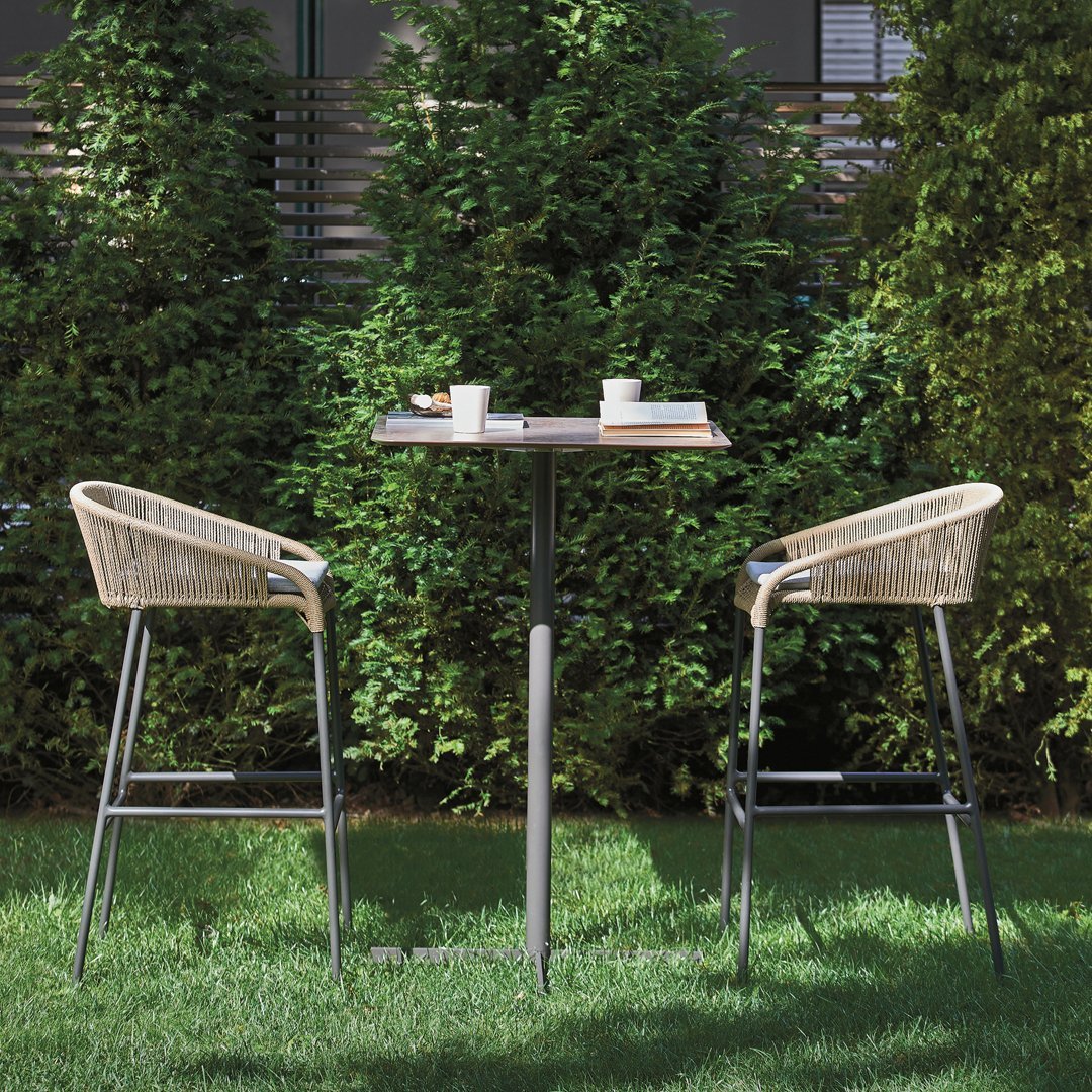 "Modern outdoor bar set featuring light beige woven seats and a sleek gray table, set against lush green foliage."