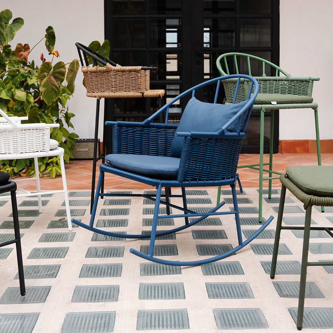 Colorful rocking chair in blue, surrounded by green, white, and brown woven chairs on a patterned gray tile floor.