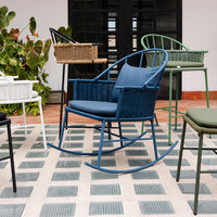 Colorful rocking chair in blue, surrounded by green, white, and brown woven chairs on a patterned gray tile floor.