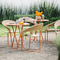 Rattan chairs and orange metal tables with intricate patterns, set against a lush green garden backdrop.