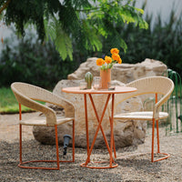 Natural beige chairs with orange accents around a round orange table, set in a green outdoor space with a stone backdrop.