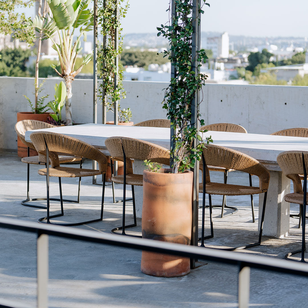 "Modern outdoor dining set with beige wicker chairs, gray concrete table, and terracotta planters under lush greenery."