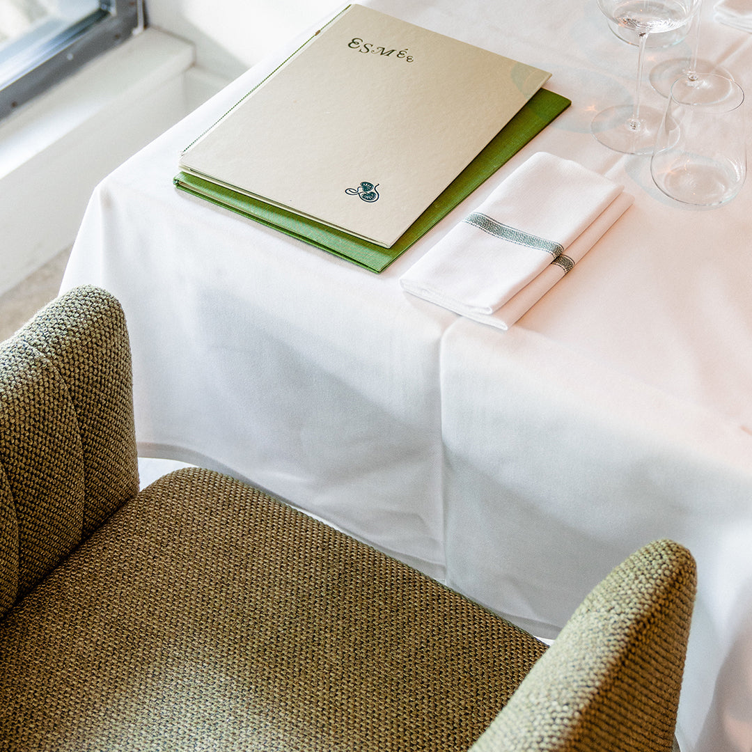 "Neutral green and beige chair with a white tablecloth and a layered green and cream menu on a dining table."